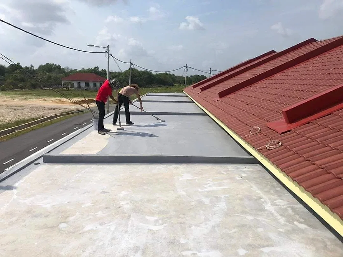 Two workers rolling waterproof coating along a flat roof beside a tiled roof
